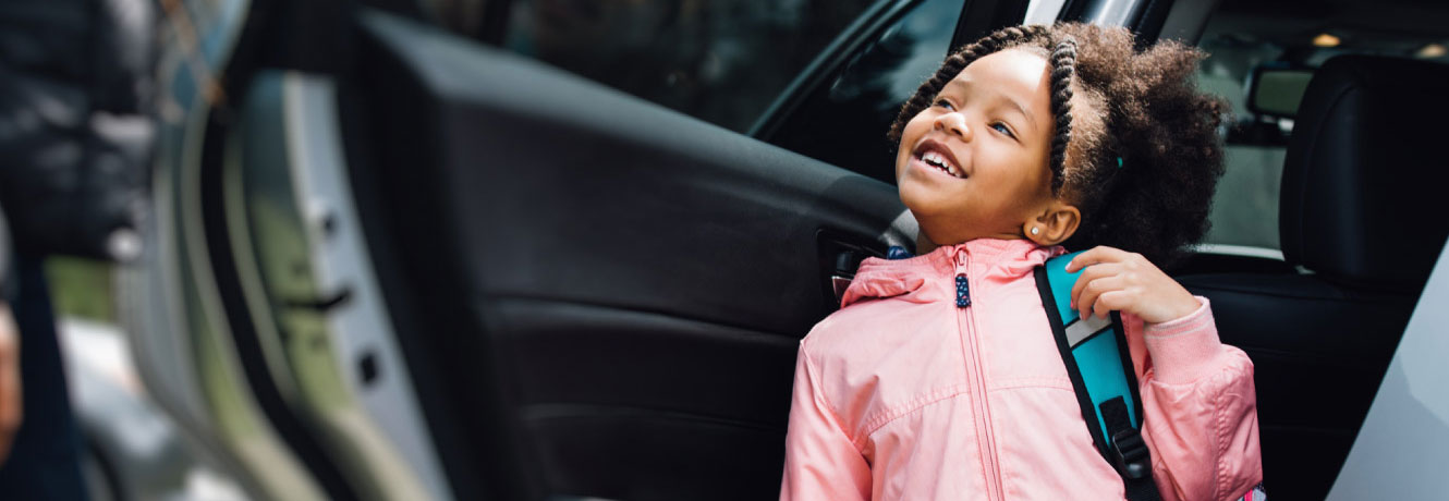 Young smiling child gets out of the back seat of a car with her back pack on.